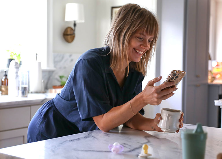 Smiling mother at home using smartphone with coffee in hand, representing convenient access to virtual care through TextCare