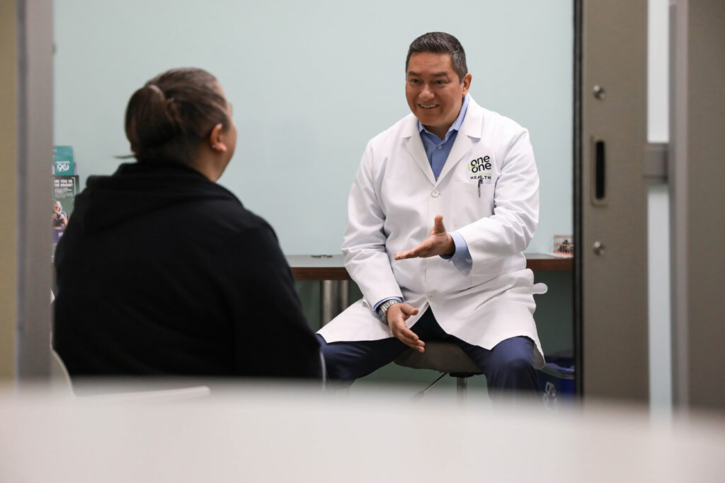 One to One Health provider in white coat talking with a patient in a private exam room, emphasizing personal and attentive care