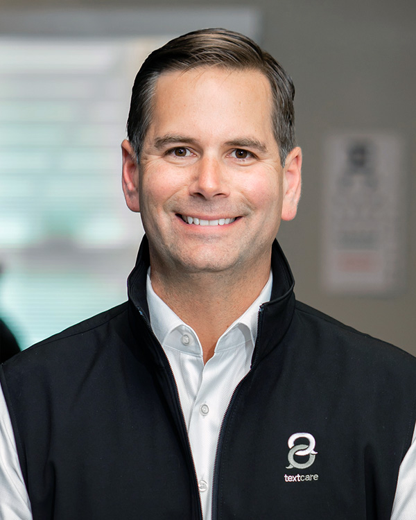Portrait of Wil Trohanis, Chief Revenue Officer at One to One Health, wearing branded TextCare vest and smiling in office setting
